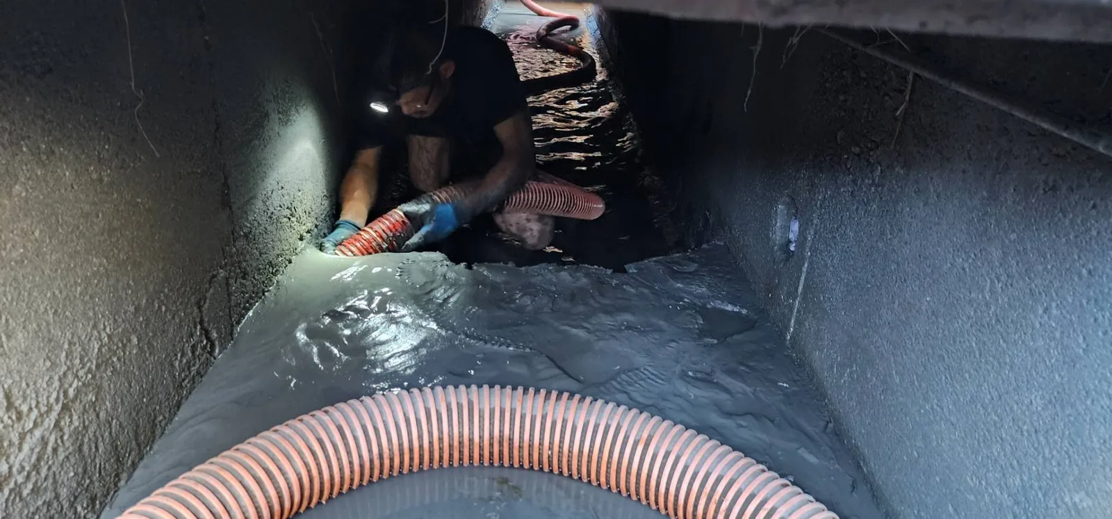 戴頭燈的師傅蹲在箱涵深處清淤，前方地面積滿灰色泥漿與吸管