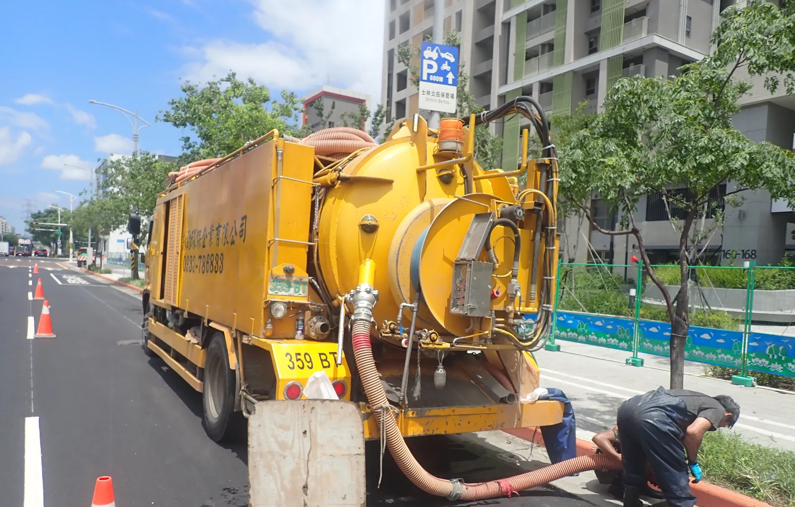 黃色清溝車停在行道樹下的路邊,旁邊是綠色圍籬與植栽